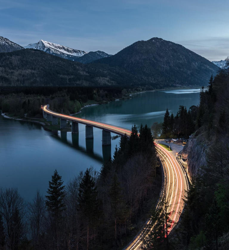 Illuminated road with bridge crossing lake with forest in foreground and mountains in background